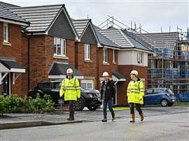 Three people in Stewart Milne-branded hi-vis jackets and hard hats walking in front of new homes
